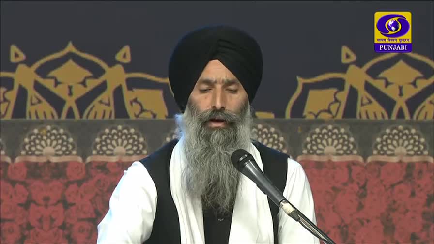 A man with a long, gray beard speaks into a microphone, his eyes closed. The DD Punjabi logo is visible, and the backdrop is a patterned design.
