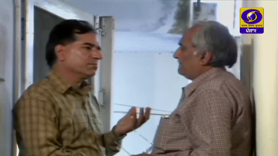 Two men stand close, talking near a doorway. The man on the left gestures with his hand as he speaks, while the other listens intently.
