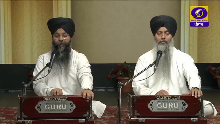 Two men, both with long white beards and black turbans, sit side-by-side, singing into microphones. They are playing harmoniums, the instruments bearing the name "GURU" and the address of a shop in Jalandhar, India, on their fronts.
