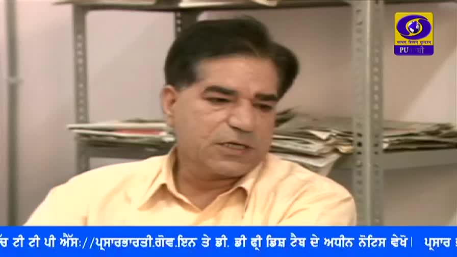 A man in a light yellow shirt is speaking, his gaze directed slightly off-camera. Behind him, a shelf holds stacks of papers, and the DD Punjabi logo is visible in the corner.
