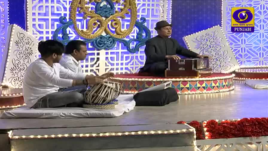 A man in a hat plays a harmonium, while two others sit nearby, their hands moving quickly over a tabla. The set, with its ornate backdrop, looks like it's from a DD Punjabi broadcast.
