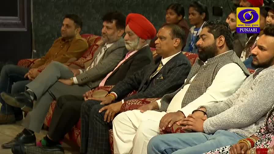 A group of men and women sit on a patterned sofa, watching something off-camera. The DD Punjabi logo is visible, and the audience appears attentive.
