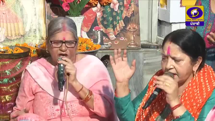 Two women are singing into microphones, their hands raised in a devotional gesture. Behind them, a vibrant religious image and a DD Punjabi logo suggest a broadcast from India.