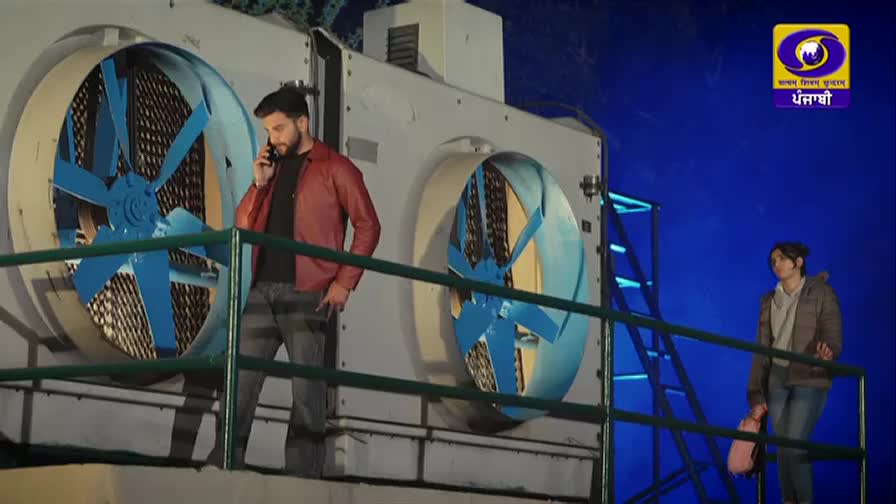 A man in a red jacket talks on his phone, leaning against a railing. A woman stands further down the walkway, holding a pink bag.