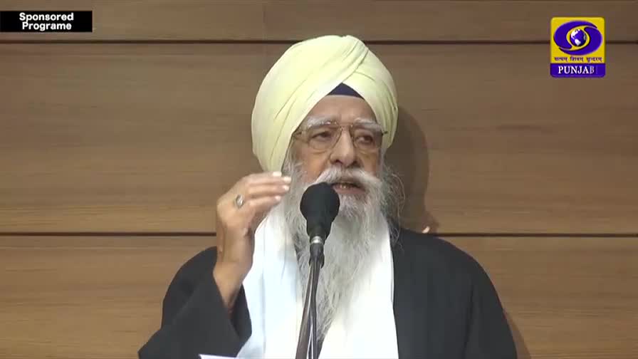 An elder Sikh man, his long white beard prominent, speaks into a microphone. He gestures with his right hand, his fingers slightly splayed, as if emphasizing a point. The DD Punjab logo is visible in the corner, indicating this is a broadcast from India.