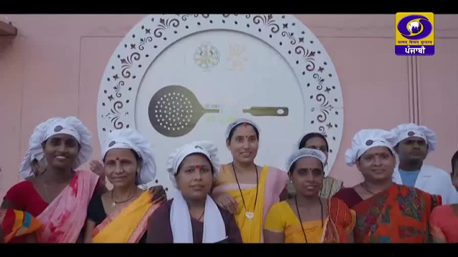 A group of women, all wearing white hairnets, stand together in front of a large circular emblem. The emblem features a stylized slotted spoon and ladle. The DD Punjabi logo is visible in the upper right corner.