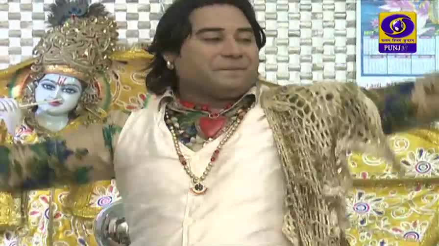 A man in a white vest and layered necklaces stands beside a statue of Krishna playing a flute. The DD Punjabi logo is visible in the background.