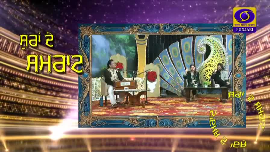 A man plays a harmonium on a stage, while two women sit on a bench to his right. The backdrop features a large, stylized peacock feather design.