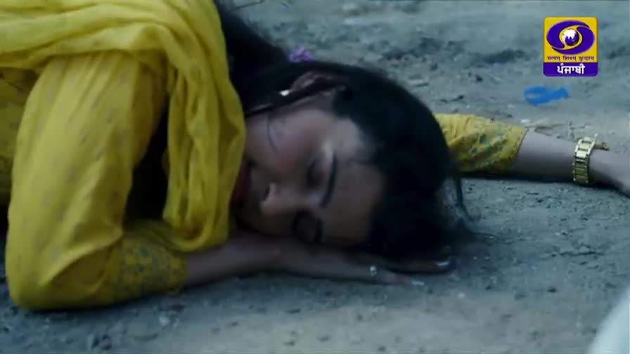A woman in a bright yellow salwar kameez lies still on the dusty ground, her face turned away. Her arm, adorned with a gold watch, stretches out beside her.