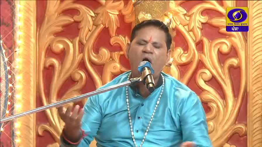 A man in a bright blue kurta sings into a microphone, his eyes closed in concentration. The ornate gold and red backdrop suggests a cultural performance, likely broadcast on DD Punjabi.