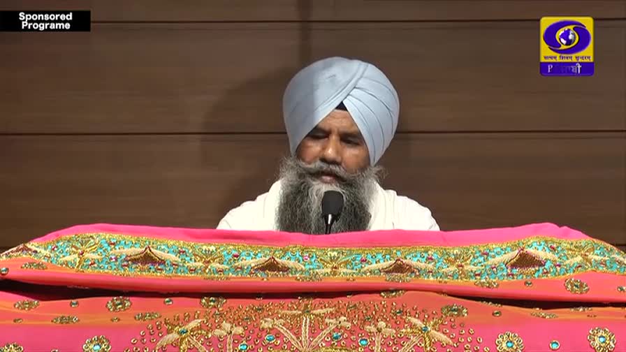 A man with a long grey beard and a light blue turban speaks into a microphone, his head bowed slightly. He's positioned behind a vibrant pink and gold embroidered cloth, likely part of a religious or cultural broadcast for DD Punjabi.