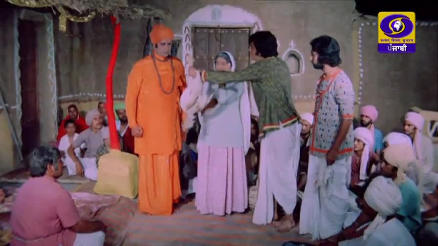 A man in an orange robe stands stoically as another man in a green kurta gently guides a woman in a light blue outfit. A crowd of people in traditional Indian attire watches from the background.