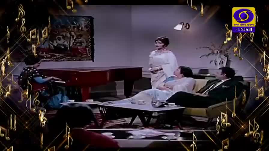 A woman in a polka-dot dress plays a grand piano while another woman, in a white sari, stands and speaks to a couple lounging on a sofa. The DD Punjabi logo is visible in the corner.