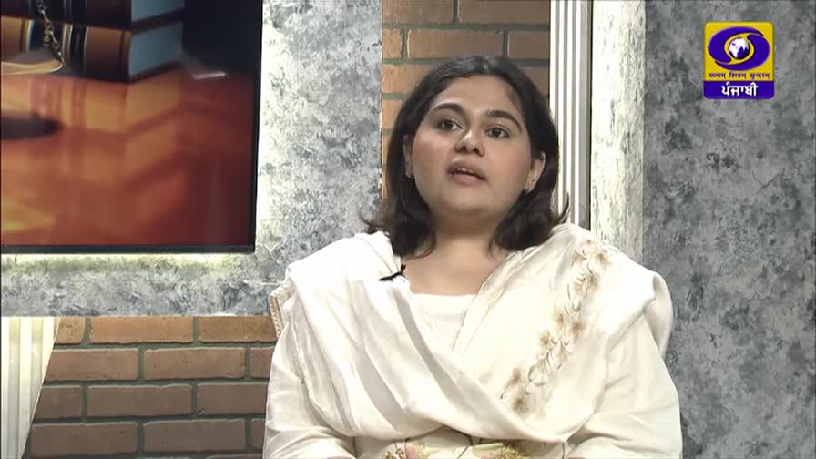 A woman in a light-colored shalwar kameez speaks on a television program, the DD Punjabi logo visible in the background. The set features a brick wall and a screen displaying stacks of books.