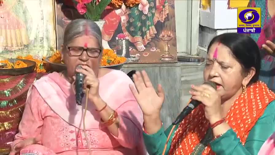 Two women are singing into microphones, their faces turned towards the audience. Behind them, a colorful depiction of deities and floral offerings suggests a religious setting.