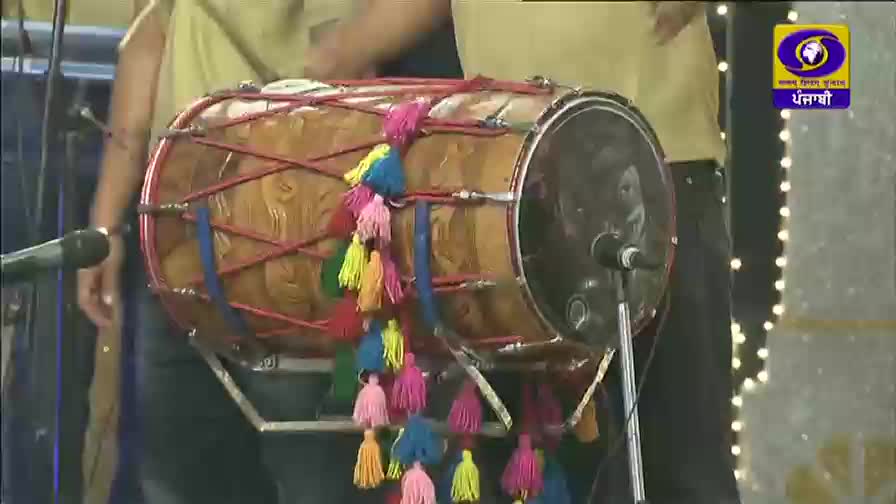 A dhol player's hands are a blur as they strike the drum, sending a cascade of colorful tassels dancing. The DD Punjabi logo is visible in the corner, hinting at a lively performance.