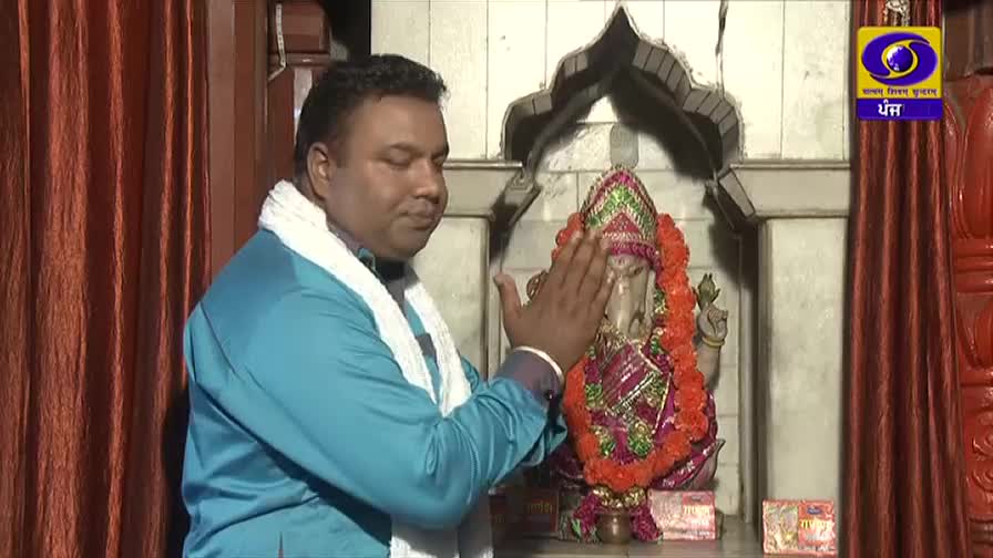 A man in a bright blue kurta and white shawl gently touches the forehead of a Ganesha idol, adorned with marigold garlands. The scene unfolds within a temple, with rich red curtains framing the background.