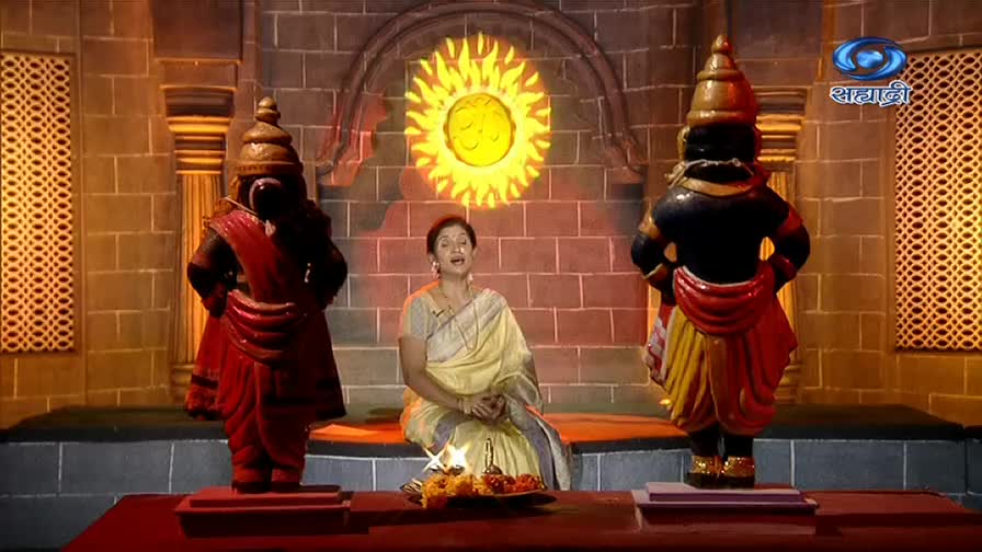 A woman in a cream sari sits center stage, speaking on DD Sahyadri, flanked by two large, ornate statues. Behind her, a painted sun radiates from the wall.
