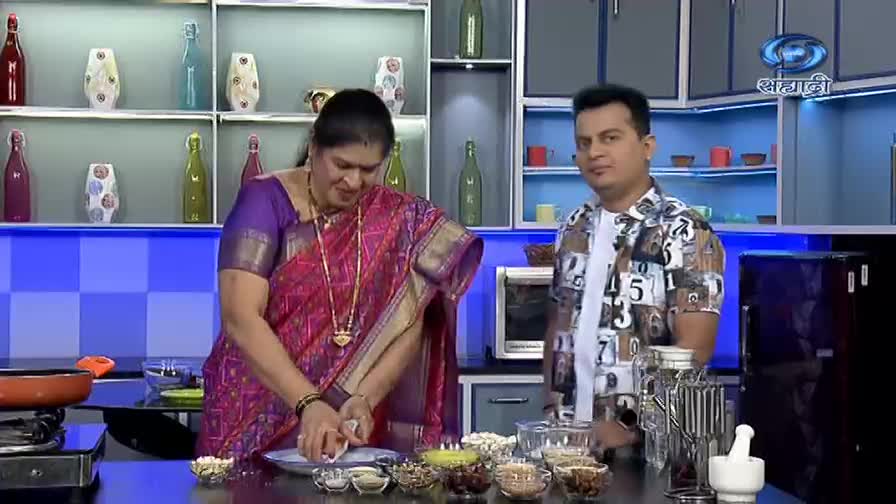 A woman in a vibrant sari is shaping something white on a plate, while a man in a patterned shirt looks on. The DD Sahyadri logo is visible in the background, suggesting a cooking segment.
