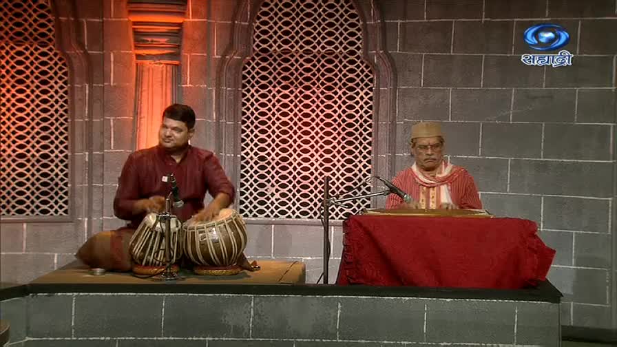 The man on the left is striking the tabla drums, his hands a blur as they move. Across from him, another man sits at a stringed instrument, ready to play on this DD Sahyadri broadcast.
