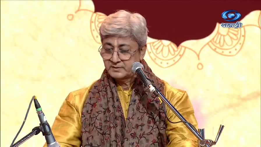 A man in a yellow kurta and patterned scarf speaks into a microphone, his glasses reflecting the stage lights. The DD Sahyadri logo sits in the upper right corner of the screen.
