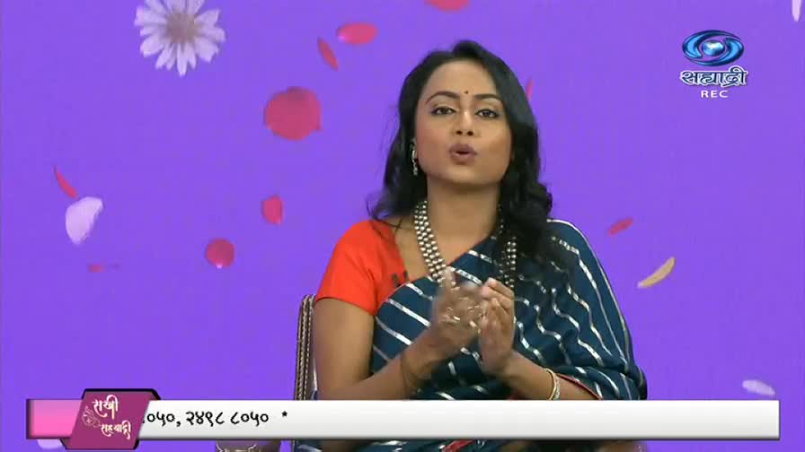 A woman in a sari speaks animatedly on a DD Sahyadri program, her hands gesturing as she talks. Rose petals and a flower float gently against a purple backdrop.
