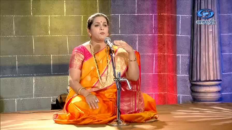 A woman in a vibrant orange sari sits cross-legged, speaking into a microphone. Her hand gestures as she addresses the audience, likely on a program from DD Sahyadri.
