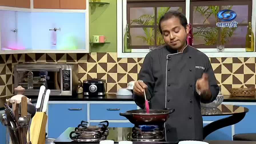 A chef, wearing a black jacket, stirs something in a pan over a blue flame. The DD Sahyadri logo is visible in the background as he gestures with his other hand.
