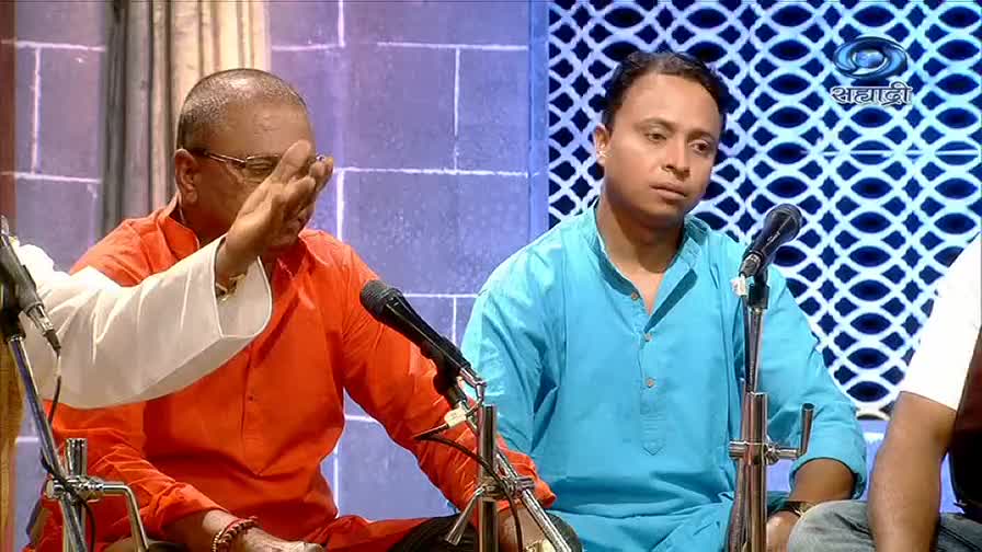 The man in orange, wearing glasses, gestures with his right hand, as if explaining something. Beside him, the man in blue looks on, both men seated with microphones in front of them, likely on a DD Sahyadri program.
