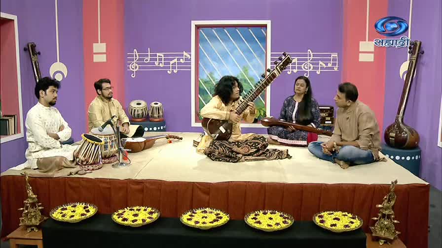 A musician with long hair strums a sitar, seated center stage on a DD Sahyadri set. Other musicians and instruments, including a tabla, are arranged around him.
