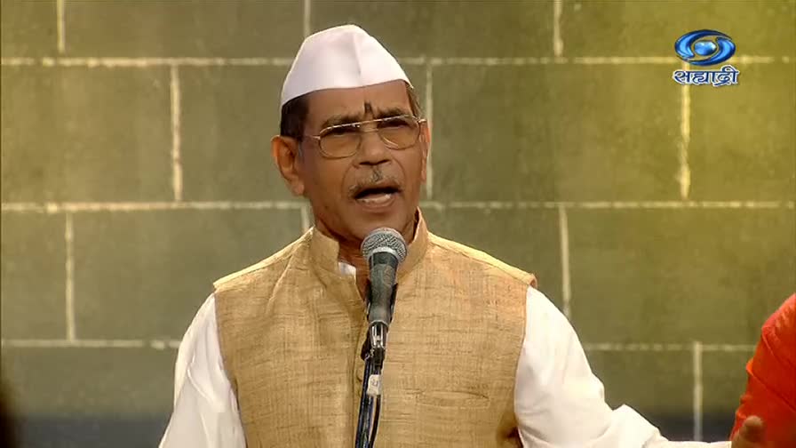 The man in the white cap and glasses speaks into a microphone, his mouth open as if mid-sentence. Behind him, the DD Sahyadri logo glows on the screen, and the background is a simple, textured wall.
