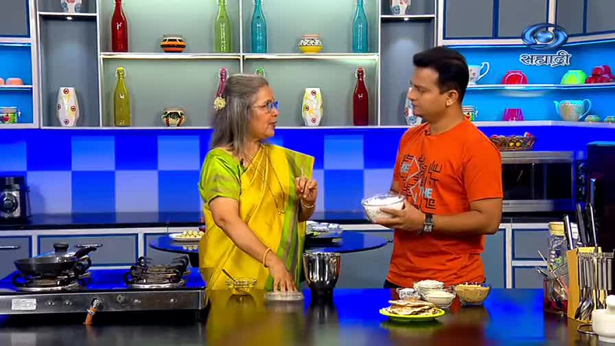 The woman in a yellow sari gestures while speaking to a man holding a bowl of white food. A cooking show on DD Sahyadri is underway, with ingredients and equipment neatly arranged on the counter.
