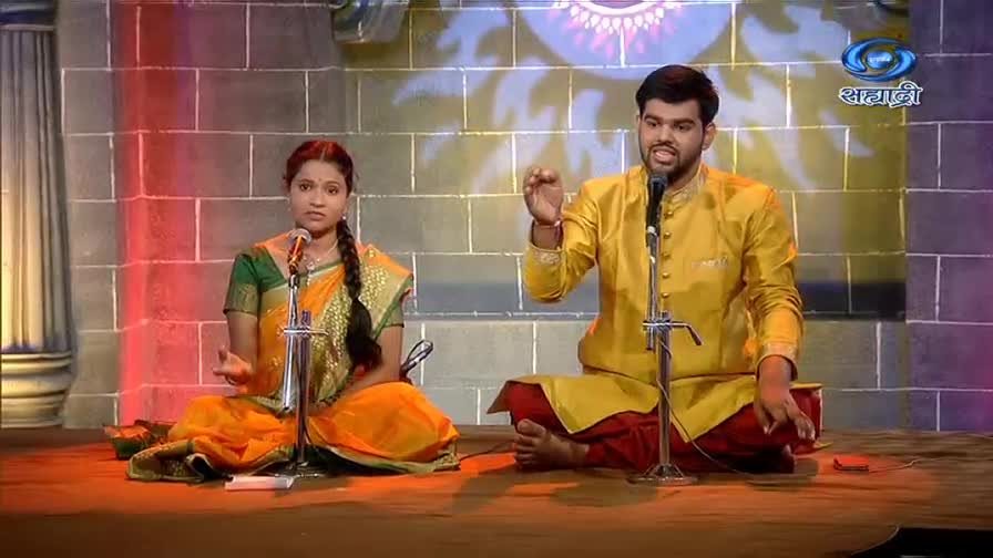 A man in a golden kurta gestures emphatically as he speaks into a microphone, while a woman in a saree sits beside him, also holding a microphone. They're on a stage, likely for a broadcast on DD Sahyadri.
