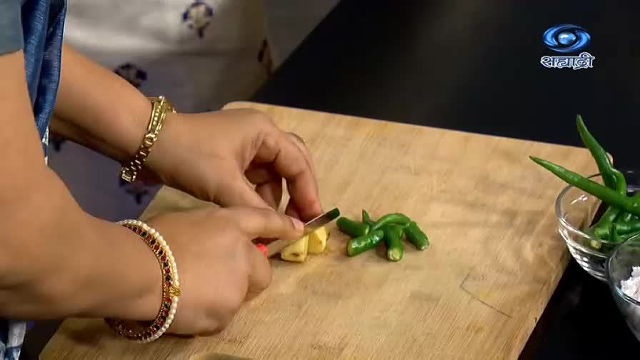 A woman, her wrists adorned with bangles, carefully slices ginger on a wooden board. Beside it, a small cluster of green chilies waits, ready for the next step in this DD Sahyadri cooking demonstration from India.
