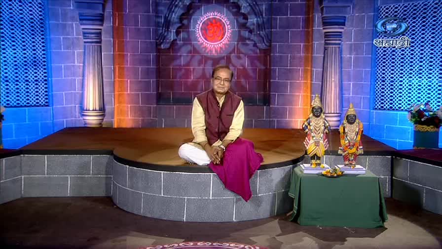 A man in a maroon vest sits cross-legged on a circular platform, a magenta cloth draped over his lap. Two small statues stand on a table covered in green fabric, lit by the studio's colorful backdrop.
