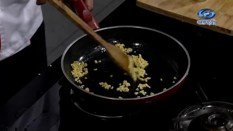 A chef, on a DD Sahyadri cooking show, stirs small, yellow pieces in a hot pan with a wooden spoon. The ingredients are glistening as they cook.
