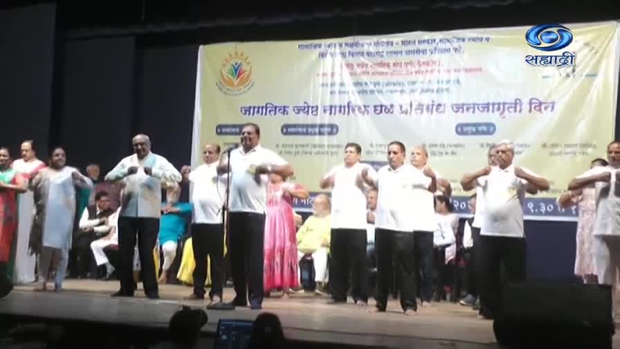 A group of people, many wearing white shirts, are on stage, doing arm exercises. Behind them, a banner is displayed, and the DD Sahyadri logo is visible on the right side.
