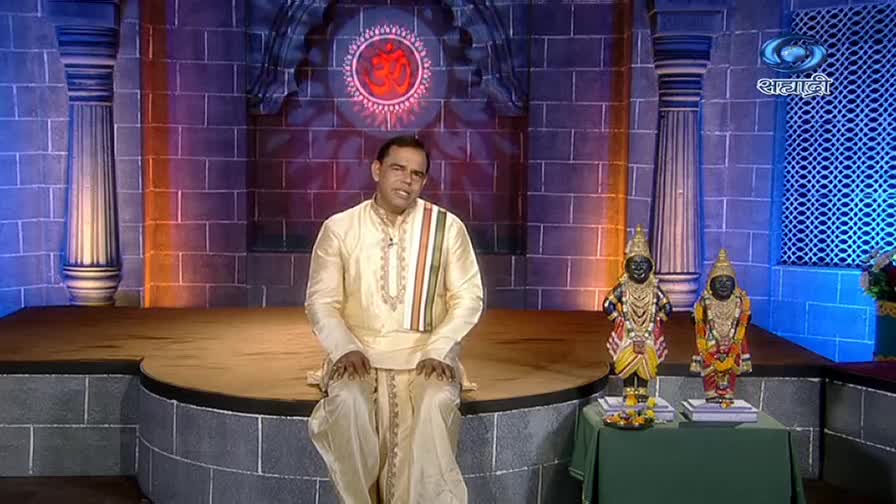 A man in traditional attire sits on a raised platform, speaking to the camera. Two ornate statues stand on a table to his right, illuminated by the studio lights.
