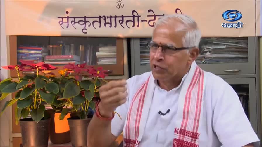 The man in the white shirt is speaking, gesturing with his right hand. Behind him, the DD Sahyadri logo and a banner with Sanskrit script are visible.
