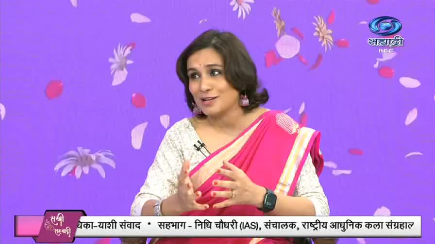 A woman in a pink sari gestures with her hands while speaking, a microphone clipped to her blouse. The backdrop is a soft purple, with scattered floral and petal designs.

