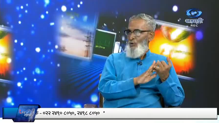 A man with a long white beard gestures with his hands, speaking on a DD Sahyadri program. The background is a swirling blue with a logo in the corner.

