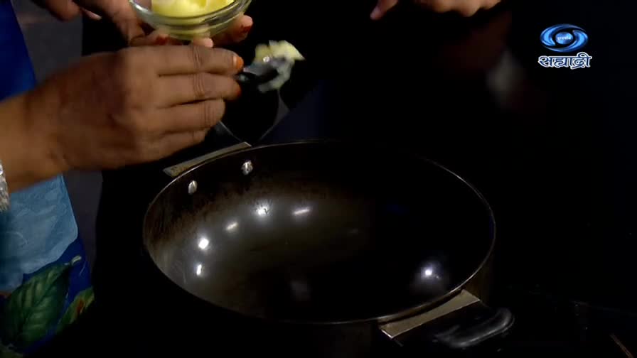 A small spoonful of ghee is being lifted from a glass bowl, ready to be added to the hot pan. The scene unfolds on a DD Sahyadri cooking show, likely in India.
