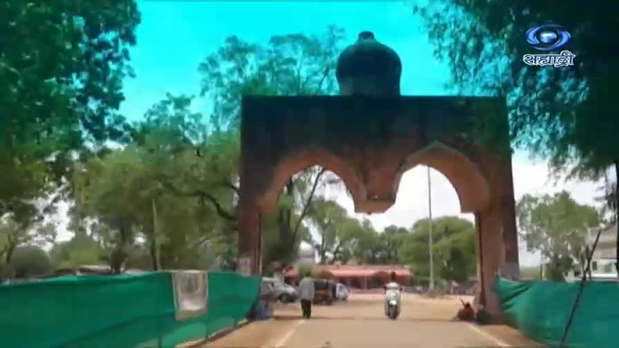 A lone scooter speeds through a large, arched gateway topped with a dome. People walk and sit along the roadside, under the shade of trees.
