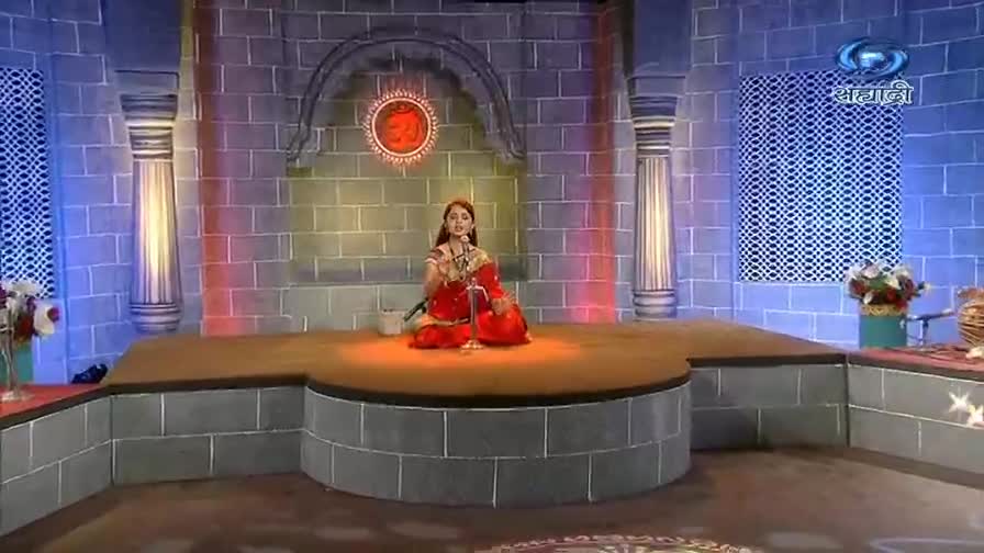 A woman in a red sari sits cross-legged on a stage, speaking into a microphone. Behind her, the DD Sahyadri logo and a decorative Om symbol are visible on the brick backdrop.
