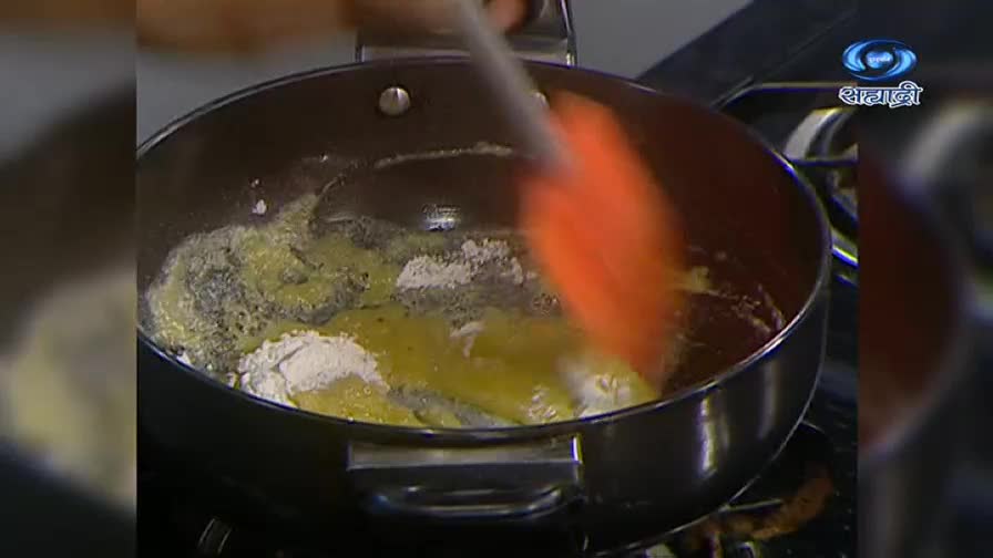 An orange spatula stirs flour into melted butter in a pan on the stove. The DD Sahyadri logo is visible in the corner, suggesting this is a cooking demonstration.
