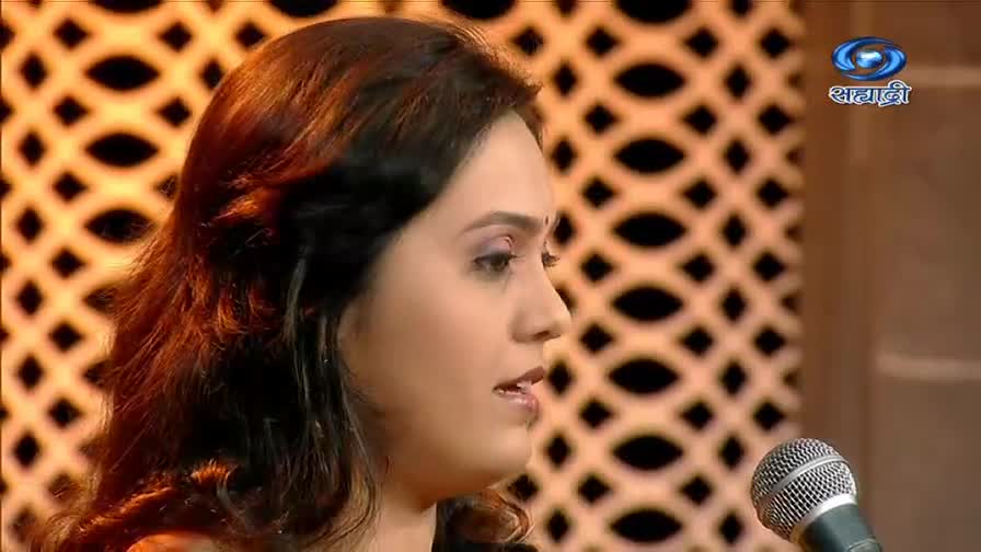 A woman with dark hair is speaking into a microphone, her gaze directed slightly to the side. Behind her, the DD Sahyadri logo is visible against a patterned backdrop.
