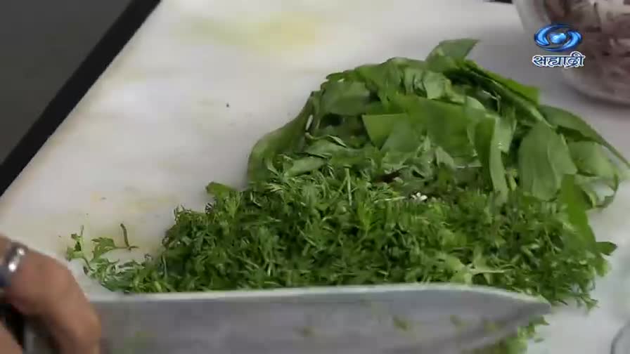 A chef is finely chopping green herbs on a white cutting board, the knife moving back and forth with precision.  DD Sahyadri's logo is visible in the corner as the cook prepares a dish.
