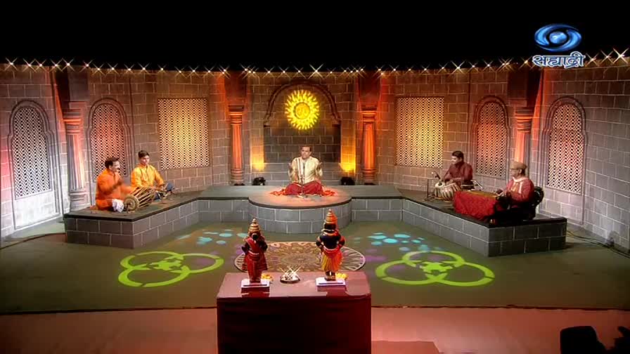 Musicians sit on raised platforms, playing instruments in a studio decorated with a sun-like emblem and stone-like walls. Two figures stand in front of the stage, and the DD Sahyadri logo is visible in the upper right corner.

