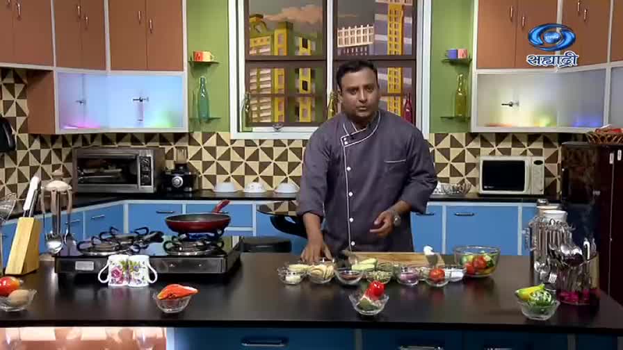 A chef in a gray jacket gestures toward ingredients arranged on a countertop, while a pan heats on a gas stove. The DD Sahyadri logo is visible in the top right corner of the kitchen set.
