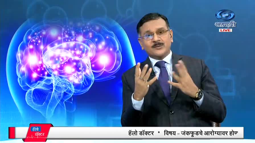 A man in a suit gestures with his hands, speaking on a DD Sahyadri program. Behind him, a glowing illustration of a brain fills the screen.
